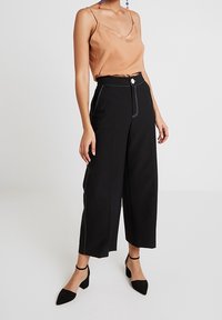 Svarta vida byxor med vit sömnad och knappstängning, kombinerat med en satängtop i mörkbeige. Modellen bär svarta pumps med spetsig tå.