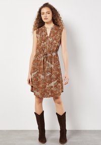 Braunes, ärmelloses Kleid mit Blumenmuster, Reißverschluss vorne und Kordelzug in der Taille, fällt über das Knie. Kombiniert mit knielangen braunen Stiefeln.