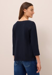 Femme aux cheveux bruns portant un haut bleu marine à manches longues et un jean bleu, tournée légèrement, montrant le dos et le profil latéral.