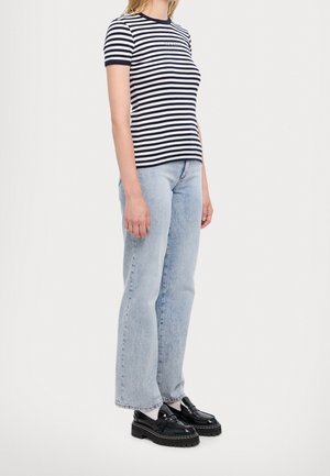 Vrouw draagt een marineblauw-wit gestreept shirt met korte mouwen, lichtblauwe jeans met rechte pijpen, witte sokken en zwarte chunky loafers, staand in zijprofiel.