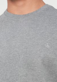 Artimas planas žmogaus, dėvinčio pilką apvalaus kaklo marškinėlį su mažuoju baltu "cK" logotipu siuvinėtame ant krūtinės.