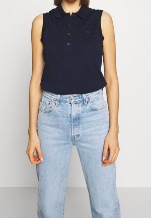 Marineblå ærmeløs polo skjorte med krave og tre knapper, parret med lyseblå højtaljede jeans. Diskret logo detaljer på skjorten.
