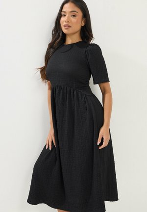 Kobieta o długich ciemnych włosach, ubrana w czarną, teksturowaną sukienkę midi z krótkimi bufkami na jasnym jednolitym tle.