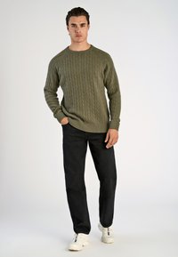 Olivengroene kabelgebreide trui met een ronde hals, gecombineerd met donkere jeans en witte sneakers, staande tegen een neutrale achtergrond.