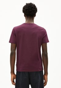 Uomo con capelli ricci che indossa una semplice maglietta a maniche corte color bordeaux e jeans scuri, visto di spalle su uno sfondo bianco.