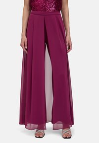 Pantalon large mauve avec superposition transparente, présentant des plis à la taille et une silhouette fluide. Assorti à des sandales à lanières argentées.