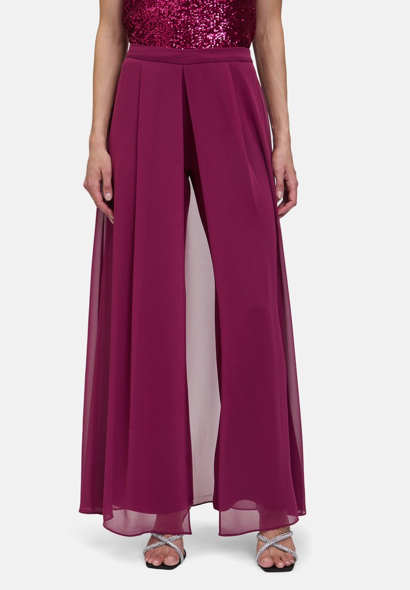 Pantalon large mauve avec superposition transparente, présentant des plis à la taille et une silhouette fluide. Assorti à des sandales à lanières argentées.