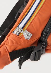 Κοντινό πλάνο πορτοκαλί υφάσματος με φερμουάρ ριγέ σε λευκό, κίτρινο και μπλε χρώμα, και ασημί μανταλάκι φερμουάρ K-Way σε μαύρο λουράκι με πλαστική πόρπη.