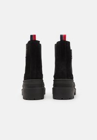 Pár černých surových platformových bot s tlustou gumovou podrážkou, elastickými bočními panely a červenými, bílými a modrými poutky vzadu.