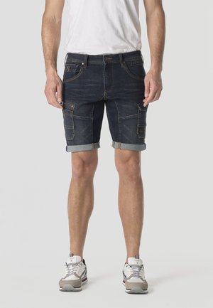 Hombre con pantalones cortos cargo de mezclilla oscura remangados, camiseta blanca y zapatillas blancas, de pie contra un fondo liso.