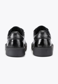 Une paire de baskets noires Kurt Geiger London avec des panneaux de talons brillants texturés et de épaisses semelles noires, vue de l'arrière sur un fond blanc.