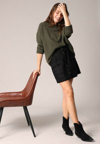 Pull en tricot vert foncé, mini jupe en suède noire avec des boutons à l'avant et des poches, associé à des bottines noires, à côté d'une chaise brune.