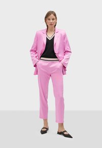 Pink blazer and trousers set, designed in smooth fabric with a tailored fit. Black V-neck top with striped accents. Black pointed flats.