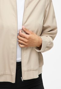 Beige bomberjakke med glidelås, ribbestrikkede mansjetter og midje, laget av glatt stoff. Modellen har den på over en hvit skjorte.