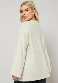 Un pull blanc duveteux avec des manches longues et décontractées, et un col rond. Texture douce avec une apparence légèrement irrégulière.