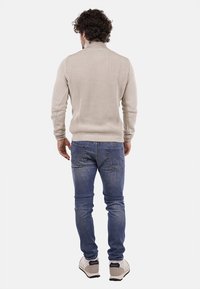 Maglione beige a coste con colletto alto, indossato con jeans blu, caratterizzato da una silhouette aderente e un tessuto testurizzato per maggiore calore.