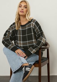 Vaalea nainen istuu ristissä jaloin puutuolilla, päällään musta, valkoinen ja ruskea ruudullinen neule, vaaleansinisiä farkkuja ja mustia lenkkareita, joissa on valkoisia raitoja.