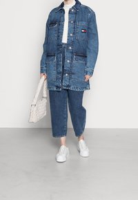 Denim jacket with large front pockets, blue color, copper buttons, paired with matching denim pants and white sneakers, holding a woven bag.