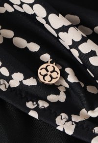 Tissu noir avec un motif floral crème, présentant un accent rond en or rose gravé avec un design floral. Texture lisse avec des détails de couture.
