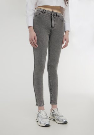 Jeans grigi elasticizzati con un design aderente, caratterizzati da cuciture a contrasto e una vita a media altezza, abbinati a scarpe da ginnastica argento metallizzato.