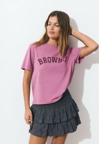 Joven mujer con cabello hasta los hombros que lleva una camiseta rosa de "BROWNIE" y una falda oscura con volantes, posando con una mano en la cabeza.