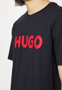 Černé bavlněné tričko s velkým červeným nápisem "HUGO" na přední straně, krátkými rukávy a kulatým výstřihem. Hladká textilie.