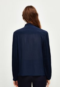 Blusa navy a maniche lunghe con dettagli plissettati. Tessuto liscio, colletto e una leggera lucentezza. Design semplice sul retro.