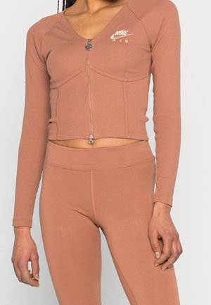 Kobieta ubrana w dopasowaną, długorękawową, krótką bluzę Nike Air w kolorze przygaszonego różu oraz dopasowane legginsy, widoczna od ramion do bioder.