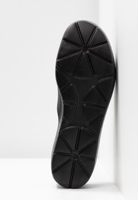 Semelle de chaussure noire présentant des motifs de traction géométriques avec des formes triangulaires segmentées et des centres circulaires, positionnée verticalement sur un fond blanc.