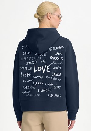 Vrouw met blond haar in een knot draagt een marineblauwe hoodie bedrukt met het woord "love" in meerdere talen op de achterkant, gecombineerd met beige broek.