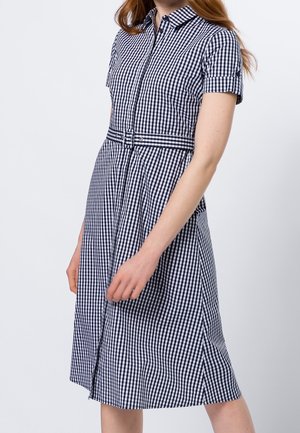 Vrouw die een knielange, korte mouw, zwart-wit geruit jurkje draagt met knopen aan de voorkant en een bijpassende riem, staand tegen een effen achtergrond.