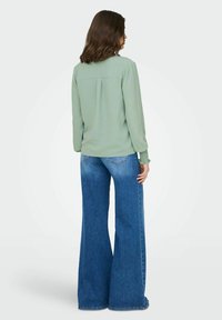 Blouse à manches longues vert clair avec un pli dans le dos, associée à un jean bleu en denim, taille haute et jambe large.