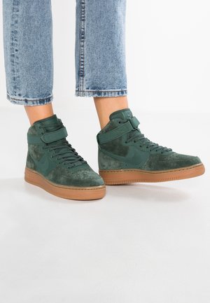 Person iført grønne ruskind høje sneakers med gummisåler og blå jeans, der er klippet over anklen, stående på en hvid overflade.