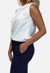 Kvinna i vit ärmlös blus med plisserad halsringning och marinblå byxor med prickar, med en hand i fickan, mot en neutral bakgrund.