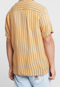 Camicetta a maniche corte di colore giallo senape con righe verticali blu navy; tessuto leggero con una vestibilità rilassata e dettagli sulla spalla posteriore.
