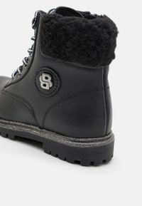 Bota de invierno negra con suela texturizada, entresuela brillante, cuello de pelo sintético, ojales de metal y un parche redondo con logo en el lateral.