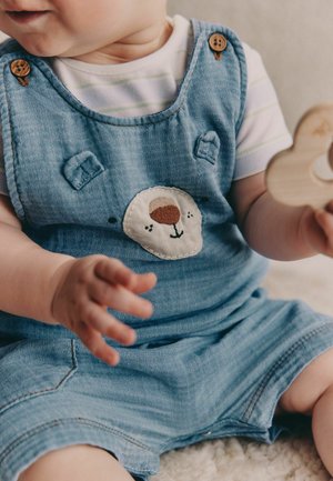 Bebeluş îmbrăcat în salopetă albastră cu patch în formă de faţă de urs, ţinând o jucărie din lemn în formă de raţă, aşezat pe o suprafaţă moale.