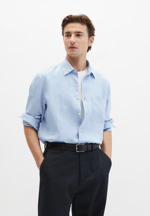 Man met bruin haar, gekleed in een lichtblauw overhemd over een wit T-shirt, zwarte riem en zwarte broek, met handen in de zakken, kijkt opzij.