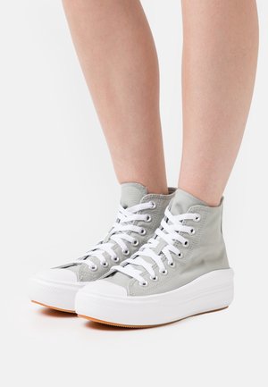 Persona con zapatillas altas de lona color gris claro, cordones blancos y suelas gruesas de goma blanca, de pie sobre un fondo blanco.