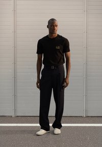 T-shirt preta de manga curta com um logótipo amarelo, combinada com calças de cintura alta azul-marinho escuro e sapatos de cor clara, sobre um fundo cinzento.