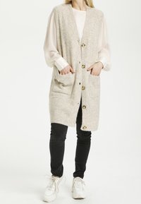 Beżowy dzianinowy kamizelka z guzikami z przodu i dwiema kieszeniami bocznymi, charakteryzująca się miękką teksturą i luźnym krojem, zestawiona z czarnymi dżinsami i białymi sneakersami.