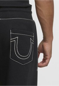 Sudadera negra con bolsillo trasero y costura blanca en contraste con un diseño en forma de U. Textura suave y cinturilla ajustada.