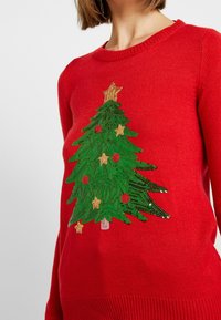Czerwony sweter dzianinowy z zielonym, ozdobionym cekinami wzorem choinki bożonarodzeniowej, z złotymi gwiazdkami i czerwonymi ozdobami. Styl z okrągłym dekoltem.
