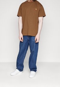 Camiseta de algodón marrón con cuello redondo y pequeño logo, combinada con jeans de mezclilla azules. Zapatillas blancas completan el atuendo.