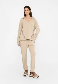 Béžová mikina s širokým výstřihem do V a bočními akcenty, doplněná vhodnými zúženými kalhotami a hnědými teniskami s designem a tkaničkami.