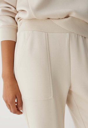 Beige sweatpants featuring a textured waistband and side pockets. The fabric is soft and slightly stretchy, with stitched details along seams.