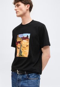 Νέος άνδρας που φοράει μαύρο μπλουζάκι με σχέδιο έρημο δρόμο και κόκκινα τριαντάφυλλα, συνδυασμένο με μπλε τζιν και μαύρη ζώνη.