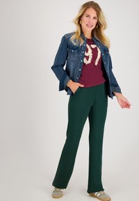 Denim overhemdjack, bordeaux t-shirt met gestructureerde opdruk, groene wijde broek en grijze sneakers. De outfit bevat zakken en gouden sieradenaccenten.