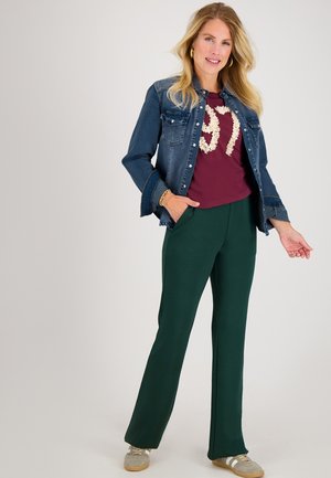 Denim overhemdjack, bordeaux t-shirt met gestructureerde opdruk, groene wijde broek en grijze sneakers. De outfit bevat zakken en gouden sieradenaccenten.