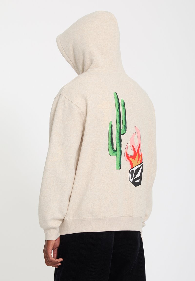 Sweat à capuche beige avec capuche relevée, arborant un motif de cactus vert et de flamme rouge-orange dans le dos, porté avec un pantalon noir.